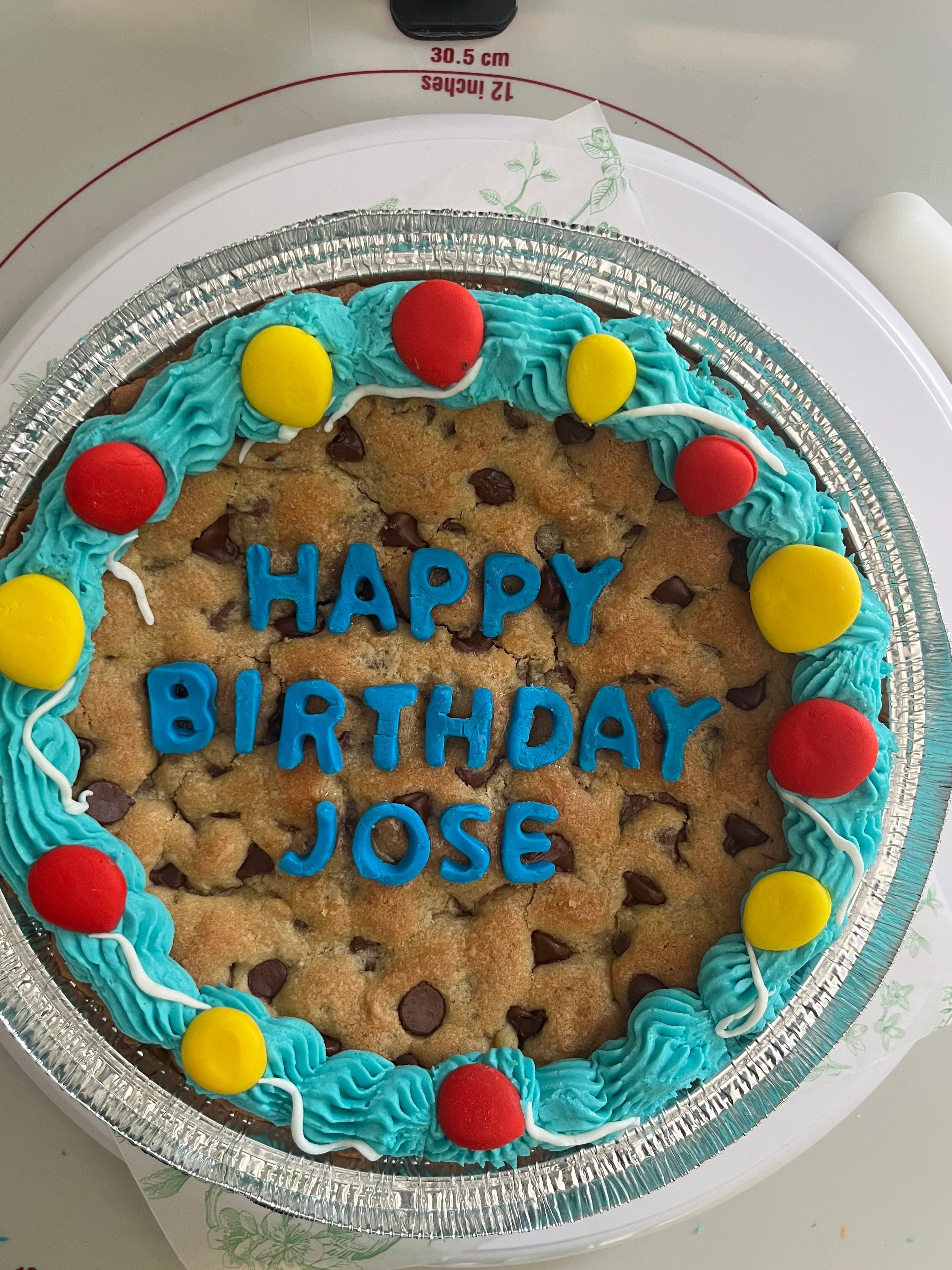 A large chocolate chip cookie cake decorated with blue frosting border, yellow and red candy-like accents, and blue lettering that reads 'HAPPY BIRTHDAY JOSE', shown on a white countertop.